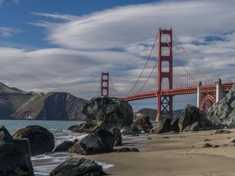 View of the Golden Gate bridge with lots of rocks on a beach and marin headlands in the distance on a partly cloudy day