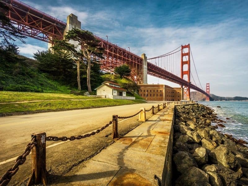 Red golden gate bridge with two buildings in the foreground on a boardwalk with rocky path