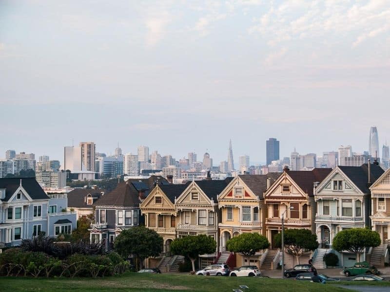 Row of pastel-painted Victorian houses going down a hill, with a patch of grass in front of it, with the San Francisco skyline behind it on a partly cloudy day.