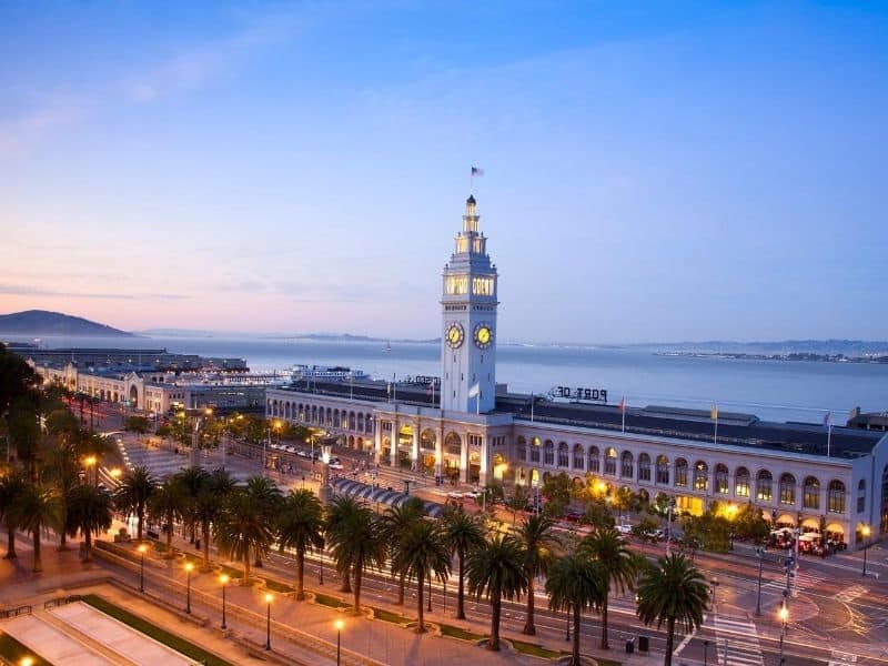 A view of the famous San Francisco ferry building, lit up at night with a clocktower and a small flag, with the Embarcadero around it.