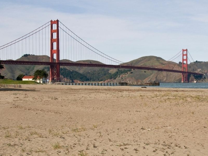 Sandy beach with bay water and red suspension bridge with hills in the distance.