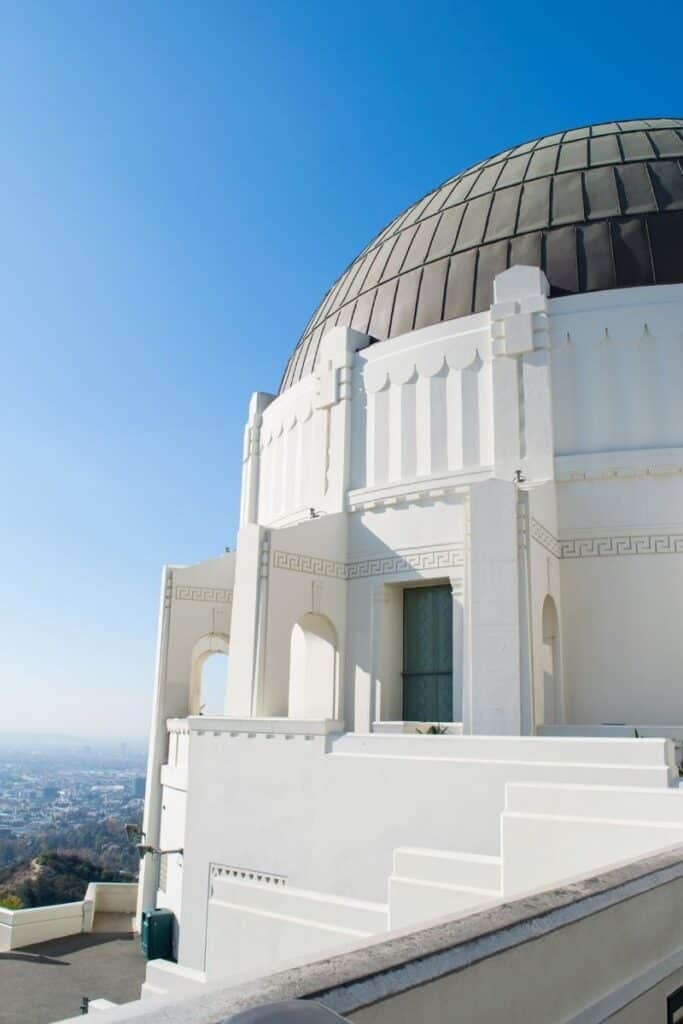 White building with gray dome roof, the Griffith Observatory in Los Angeles, with view of city in distant background below.