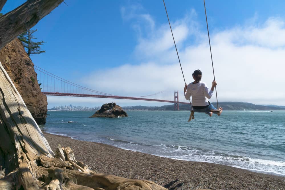 Woman on a swing on a beach with a view of the Golden Gate Bridge a red bridge going over a turquoise bay