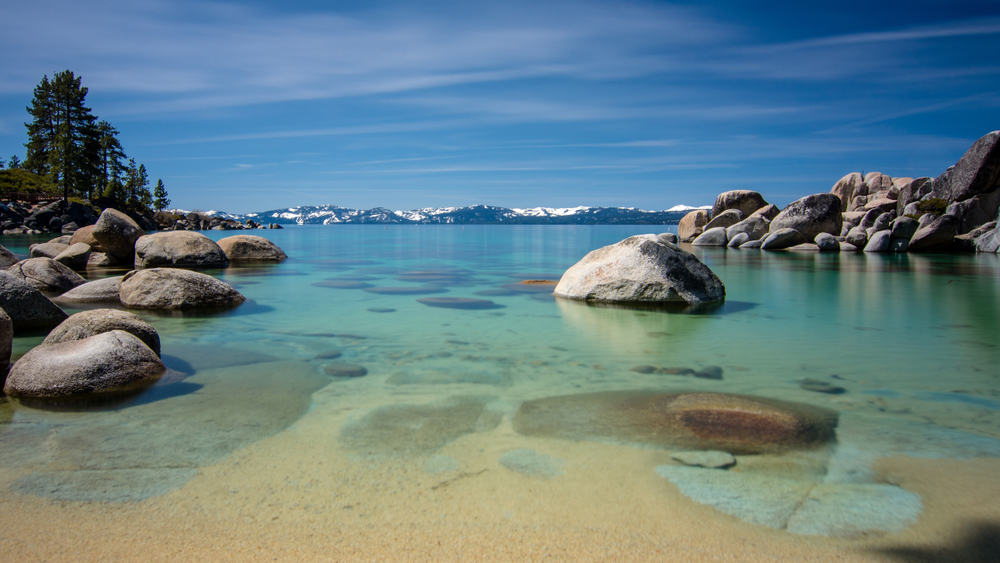 blue transparent water, rocky shore and snow on tops of background mountains on a blue sky day with few clouds