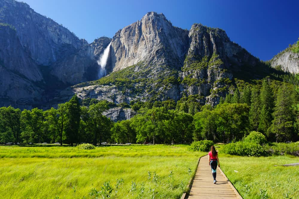 Woman in red shirt and athletic leggings walking on wooden boardwalk in the middle of a grassy meadow with a waterfall in distant background.