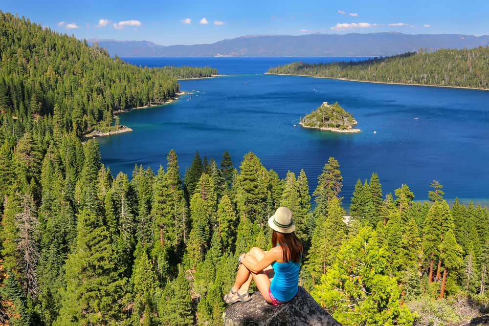 A female redheaded hiker in Tahoe who has hiked to a stunning view of Emerald Bay, where you can see cerulean blue water surrounded by pine trees and one tiny lone island in the water.