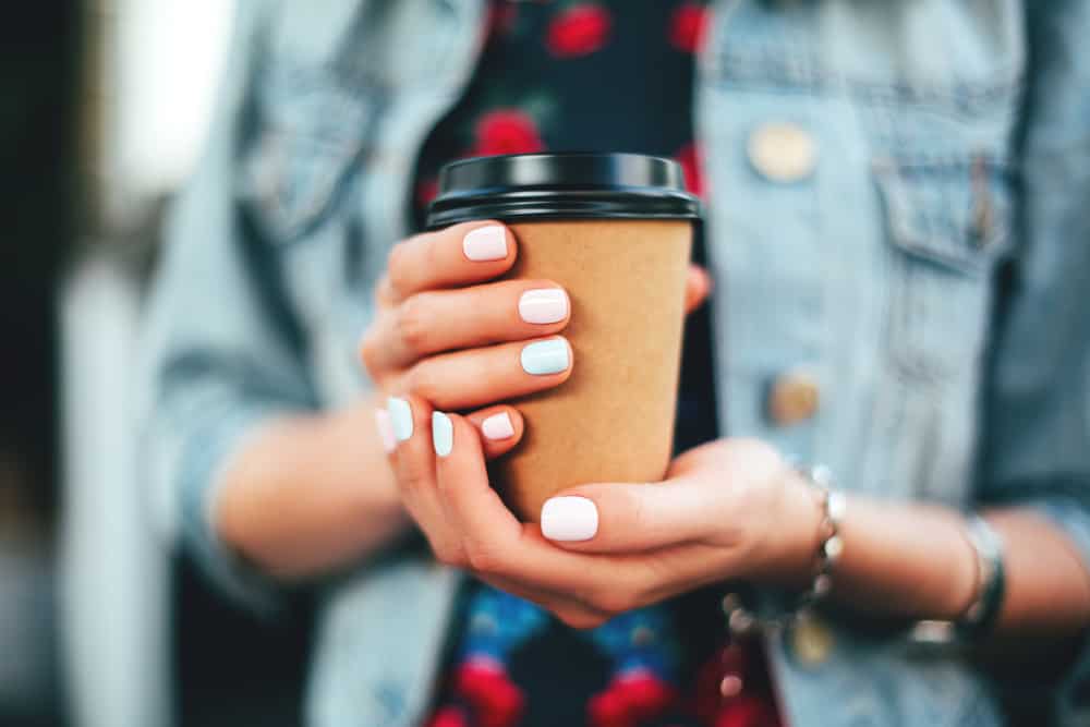 Pastel manicured hands holding a paper cup of coffee wearing a denim jacket and a flower print colorful dress.