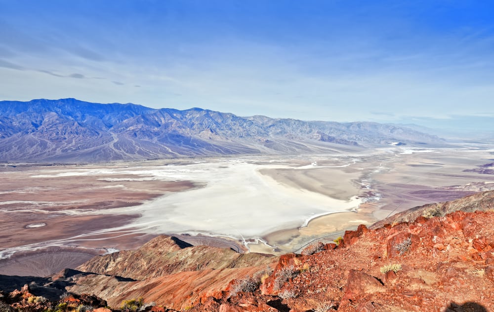 View of white salt flats in the middle of the valley floor of Death Valley's lowest point, seen from above, with red, purple, and brown rocky landscape in the desert.