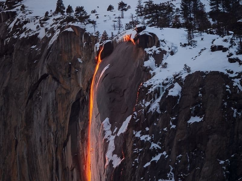 Snow on the ground of a cliff with a waterfall streaming over the edge glowing a brilliant orange, almost like. a stream of lava, from the setting sun lighting it up from behind