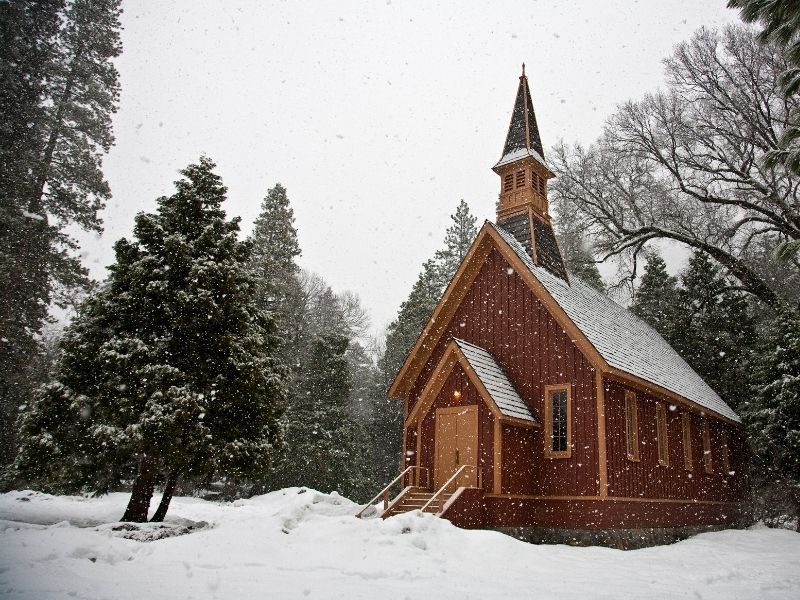 Famous Yosemite chapel (a small reddish-brown church) in the lightly falling snow with trees covered in snow and white sky.