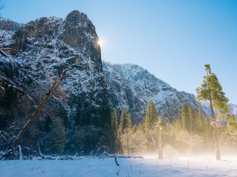 Sunburst from behind massive granite cliff, causing half of the photo to be lit up in a morning sun glow and the other half to be in shadow. Snow covered landscape.