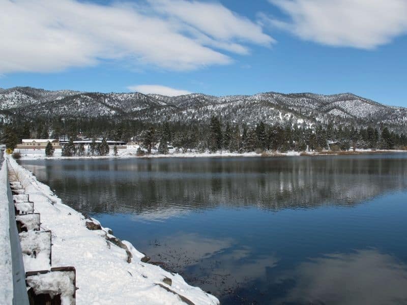 Still lake with snow on the side of lake and on the mountains in background with blue sky.