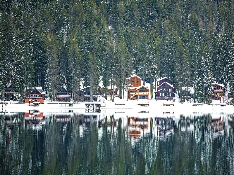 A view of the snow-covered cabins on Donner Lake in winter reflecting in the water with pine trees in the background.