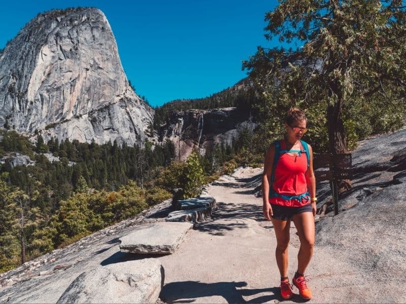 Woman in red shirt and sunglasses and shorts with turquoise backpack and red shoes on a Yosemite trail.