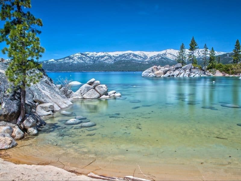 Caribbean-blue water ranging from light green to deep blue in Tahoe campground in Northern California, with snow on mountains in background and pine trees and rocks on lake shore.