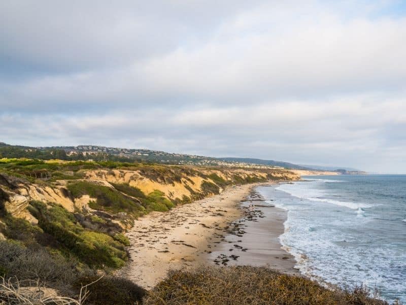 Tan cliff bluffs leading to rock-strewn beach with ocean waves and an overcast sky.