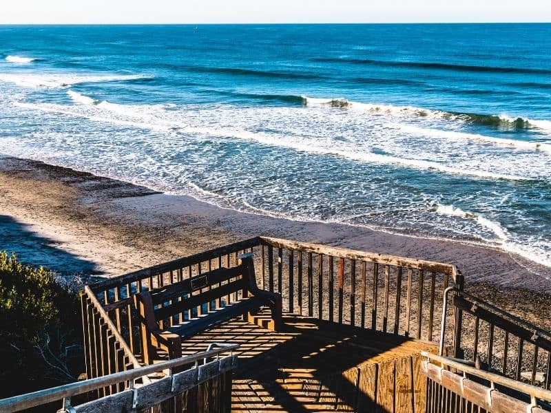 Wooden curving staircase leading to beach with cerulean blue ocean waves lapping calming on shore.