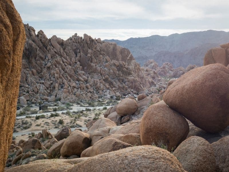 Giant boulders in foreground in shot of Indian Cove, one of the most famous Southern California campgrounds in Joshua Tree, with desert landscape behind it and more rocks.