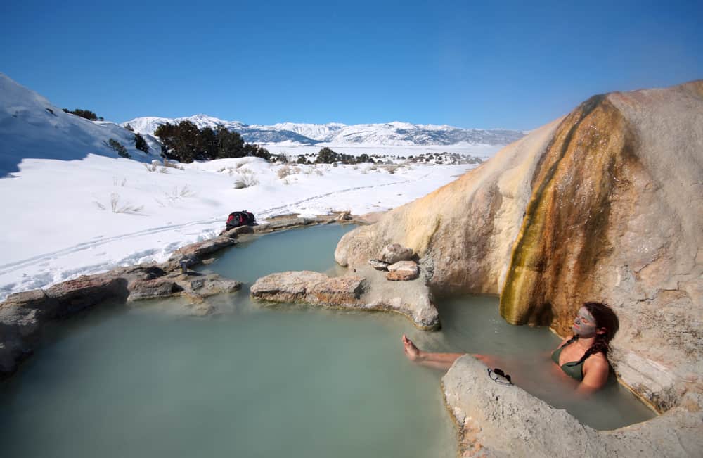 Woman with healing mud mask on her face in Travertine Hot Spring with murky light green water and snow covered landscape around her.