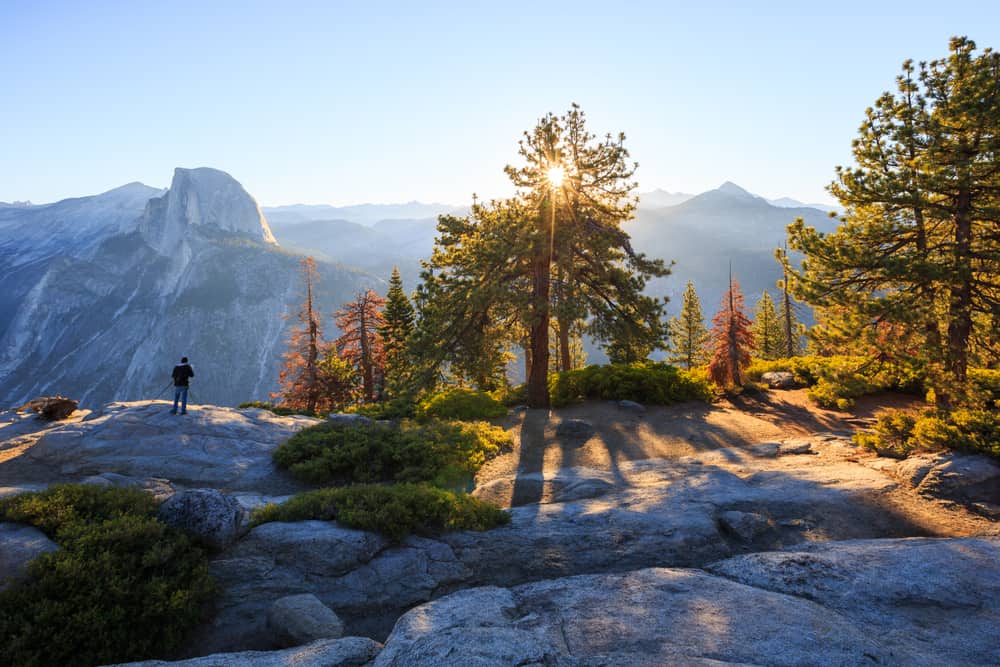 Sunburst through a tree with rocks in shadow with one man standing on cliff edge with Half Dome granite peak in background.