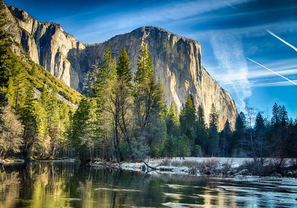 Blue sky with streaky clouds and jet streams with trees and granite cliff faces and a still river reflecting the granite.