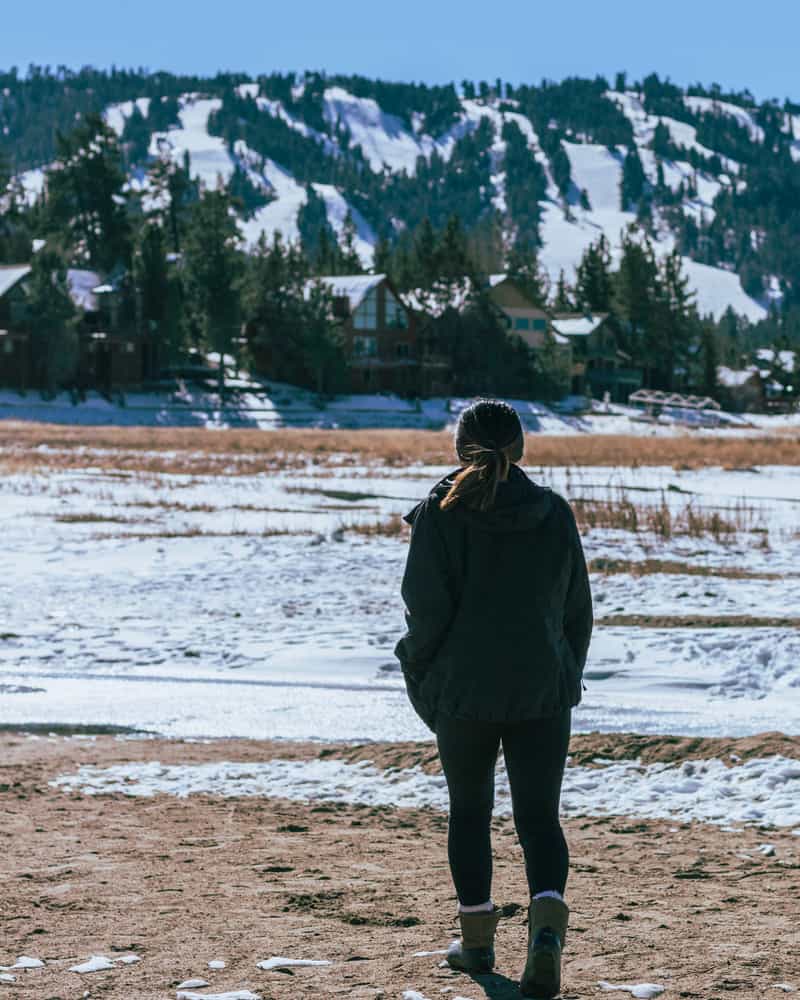 A woman dressed in black with snow boots and a ponytail with her back turned to camera looking at snowy landscape in Big Bear