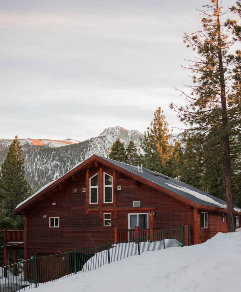 Red toned cabin in snow with view of snow-covered mountain in background with lots of pine trees and afternoon sunlight on the trees.