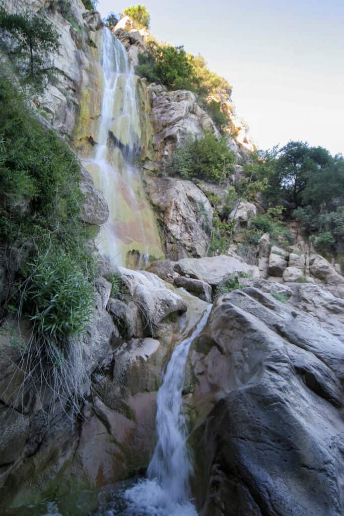 A large vertical image of the Tangerine Fall waterfall, which cascades over green and orange-tinted rocks on a sunny day.