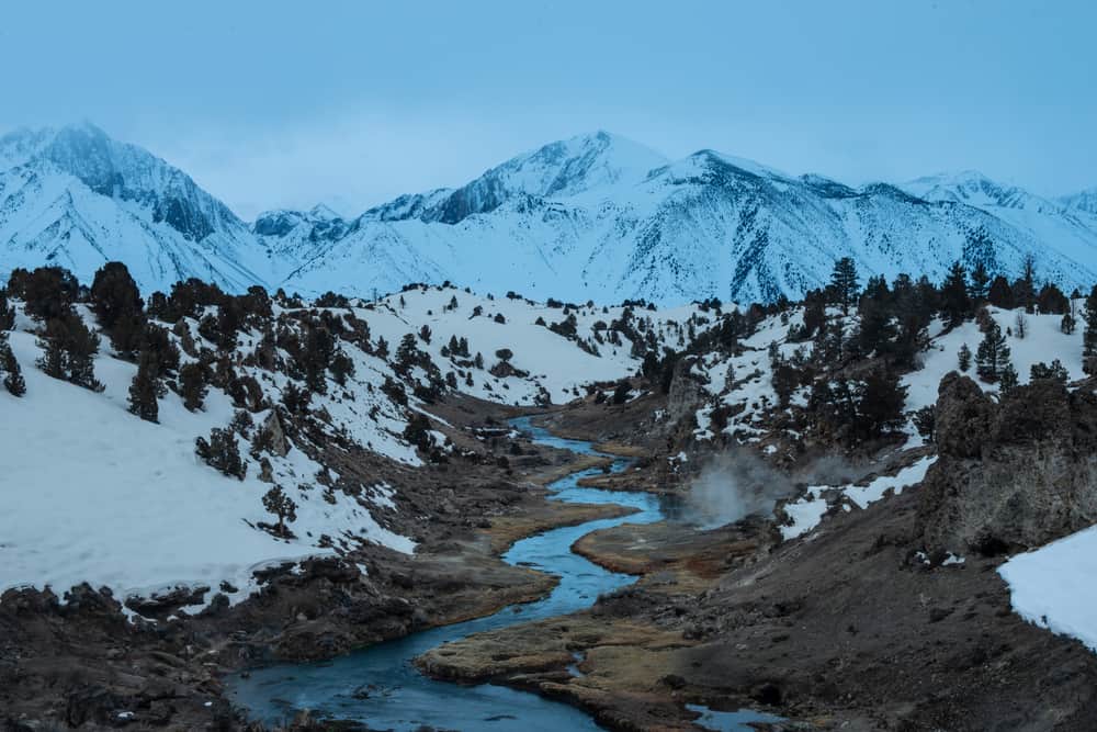 Snow-covered landscape except for creek running through a valley emitting steam from the geothermal waters. Dark cloudy sky.