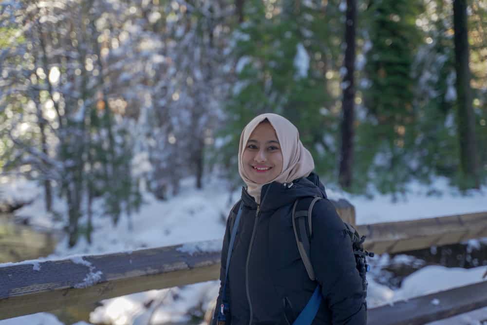 Young Muslim woman in hijab smiling at camera wearing winter clothing with snow and trees in the background while visiting Yosemite in winter.