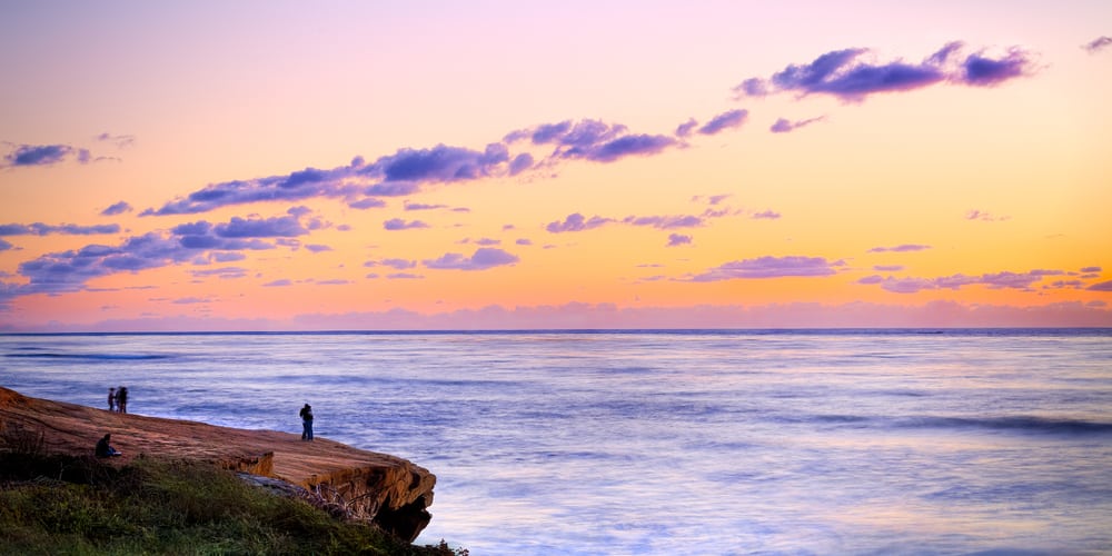 Orange sky with purple clouds and lavender-blue sea with people on cliff edge