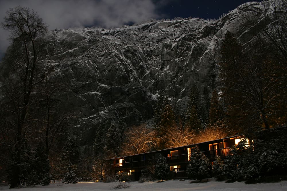 Mountain lodge lit up with orange glow at night with background of snow-covered granite rock cliffs.