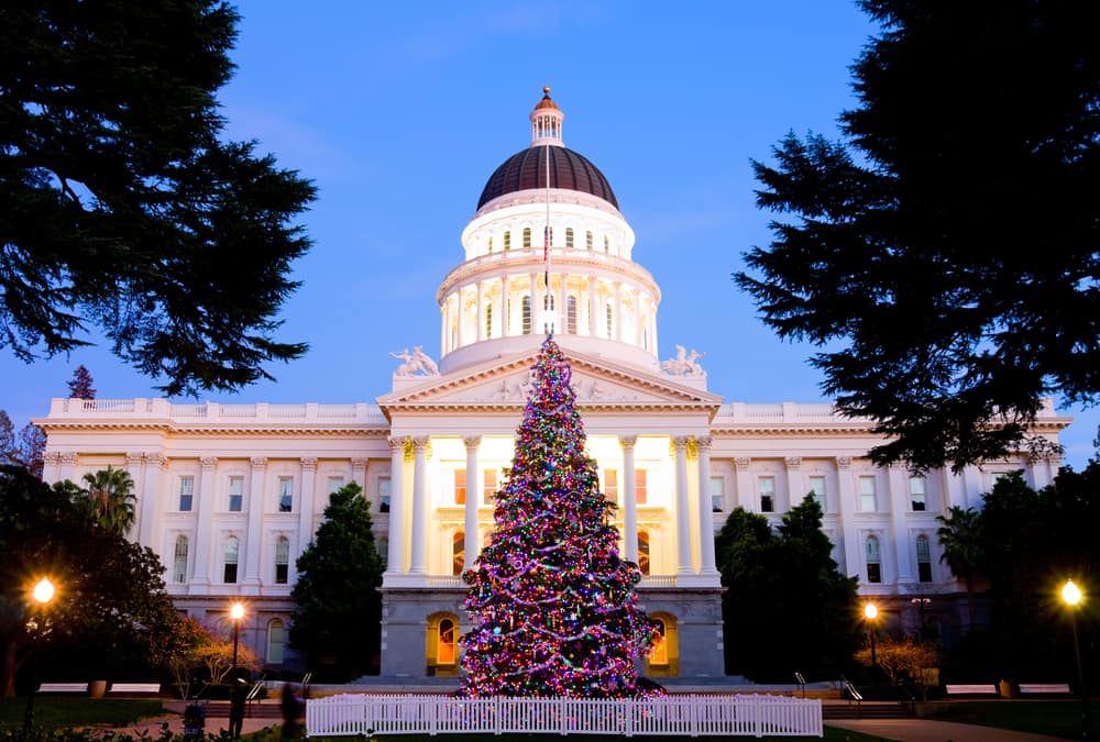 A multicolor lit up Christmas tree in front of the bright white state capitol building at twilight.