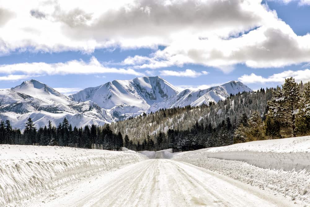 Plowed highway with snow banks on either side on a hill leading towards focal point of Mammoth Mountain and other mountains.