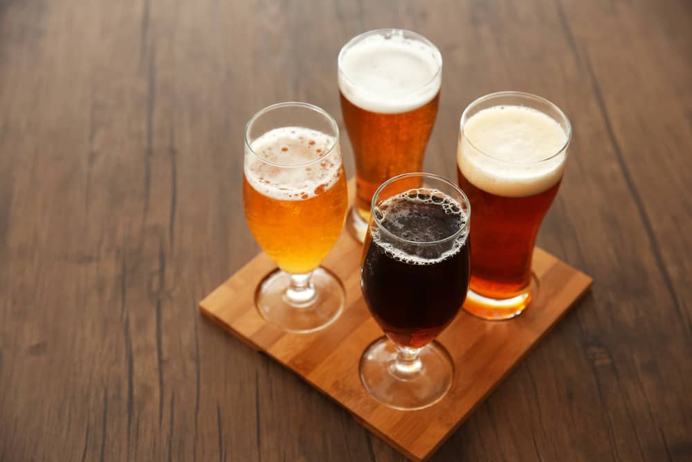 Four different kinds of beer in different colors arranged in a square on a tasting flight block of wood.