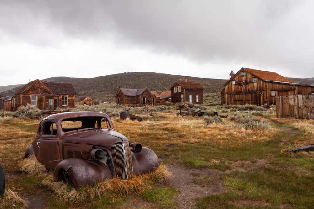 A ghost town surrounded by dilapidated homes long forgotten and a rusted car, the nature reclaiming the ghost town.