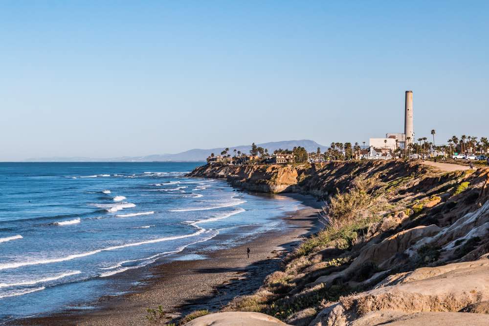 Cliffs of the Carlsbad bluffs, a beach with waves lapping onto the shore, blue sky and tall palm trees and buildings in the background.