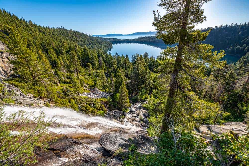 Waterfall cascading down a hill in Lake Tahoe, with a view of the glassy still Emerald Bay in the distance and Lake Tahoe further away, with tons of pine trees around.