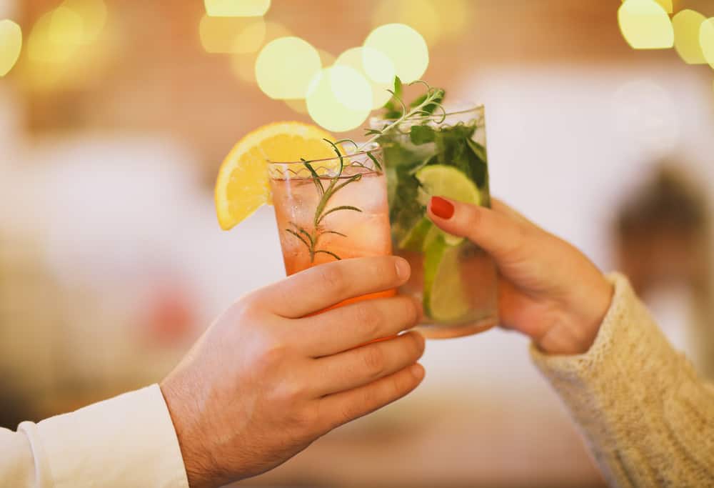 Male and female hand cheering their drink: one a pink drink with grapefruit, the other a mojito drink with mint and lime.