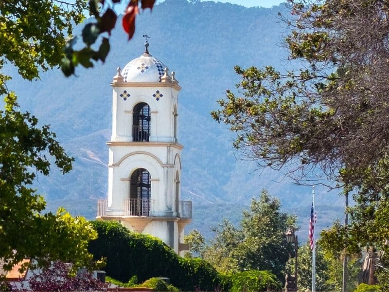 A church steeple with a background of mountains and trees and an American flag in the town of Ojai, a popular day trip from Santa Barbara