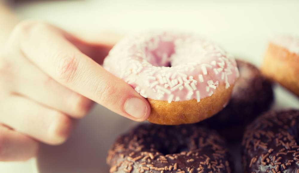 Hand holding a donut with strawberry frosting with white sprinkles and chocolate frosted donuts below it.