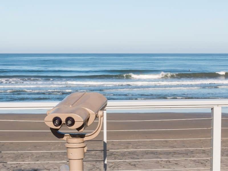 A handrail on a beach boardwalk with a white set of binoculars for whale watching standing in view, with blue water and waves.