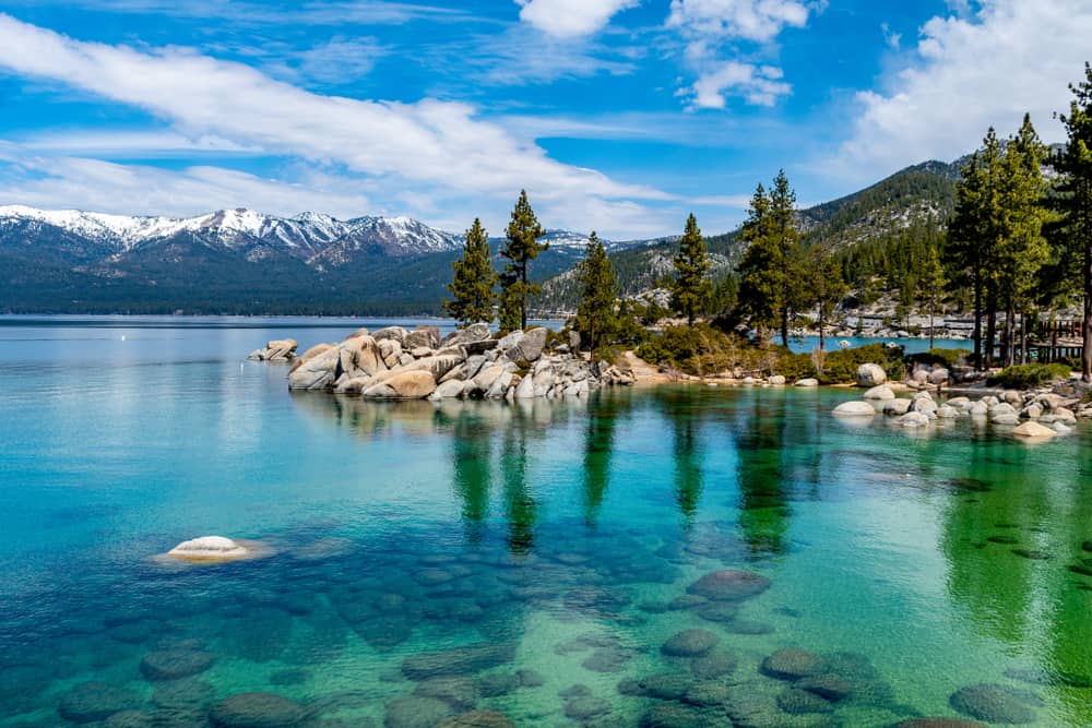 Clear water in Lake Tahoe that is turquoise, almost green, with rocks and pine trees on shore and snow-capped mountains in the distance.