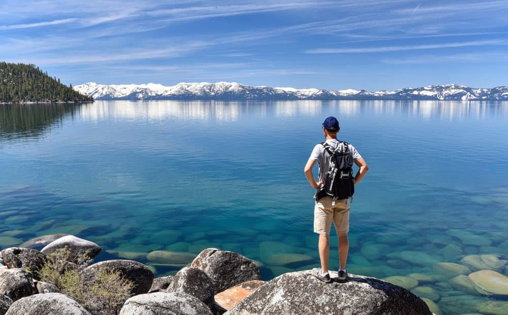 Caucasian man hiking in Lake Tahoe during the summer, looking at the turquoise water and seeing snow-covered mountains in the distance.