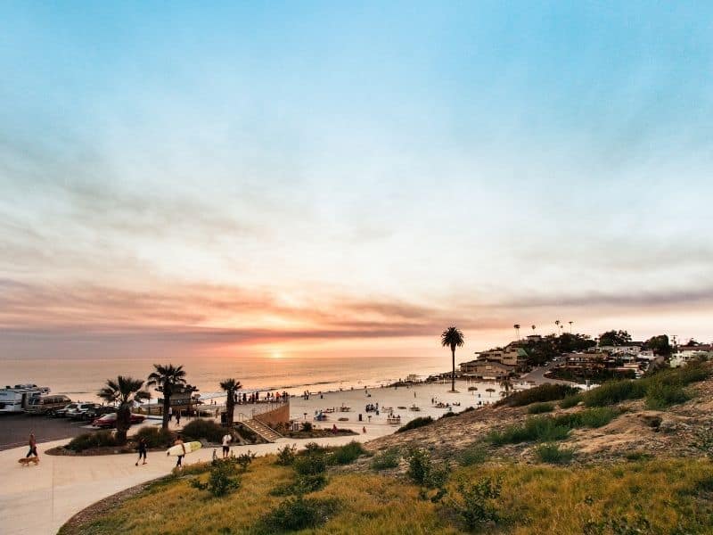 A wide angle view of Moonlight Beach in Encinitas, with people on skateboards and on the beach with palm trees and a setting sun.