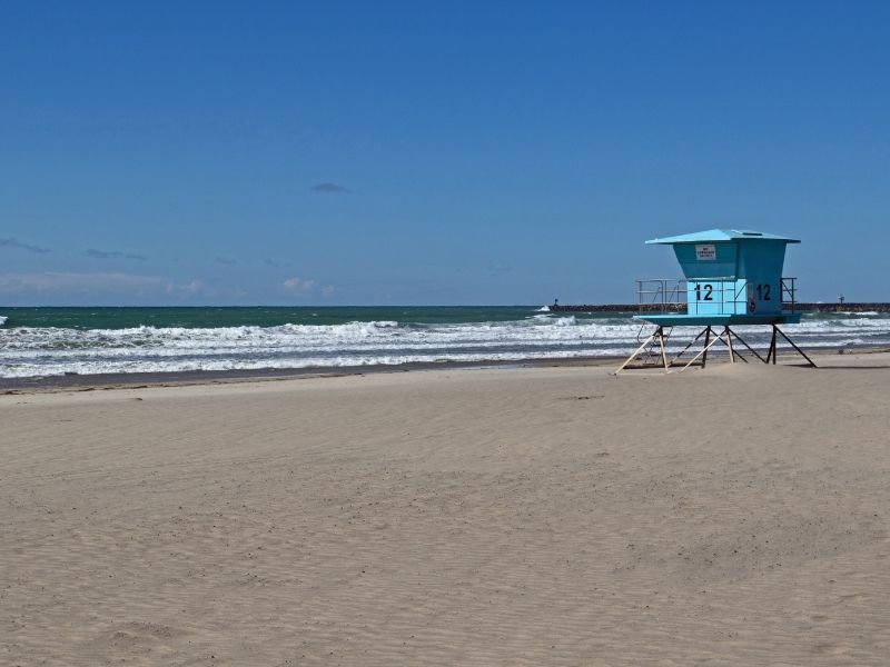 A blue lifeguard box like you see in beaches in Southern California on a sandy beach with turquoise water on a sunny day.