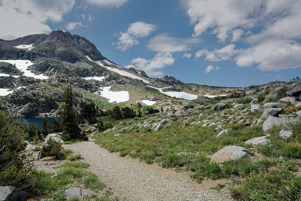 A gravely hiking path leading from a lake, the path connects Winnemuca Lake above with Frog Lake and other destinations along the Pacific Crest Trail. View of clouds in the sky, melting snowpack, and grassy trail.