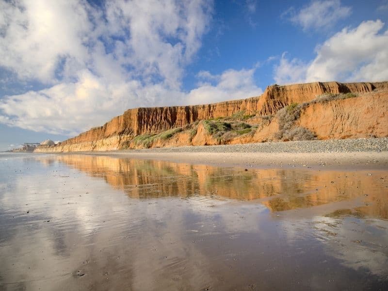 Orange-tan cliffs on a beach reflecting in the wet sand with a partly cloudy sky in San Onofre beach in San Diego County.