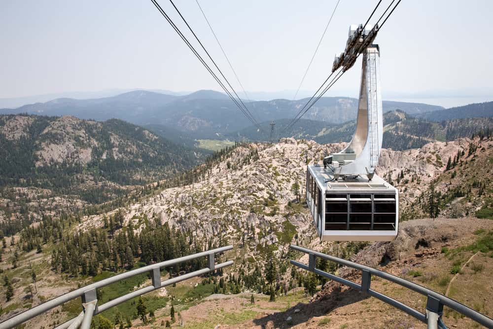 Aerial tramway car hoving over an alpine landscape of rocks, mountains, and pine trees.