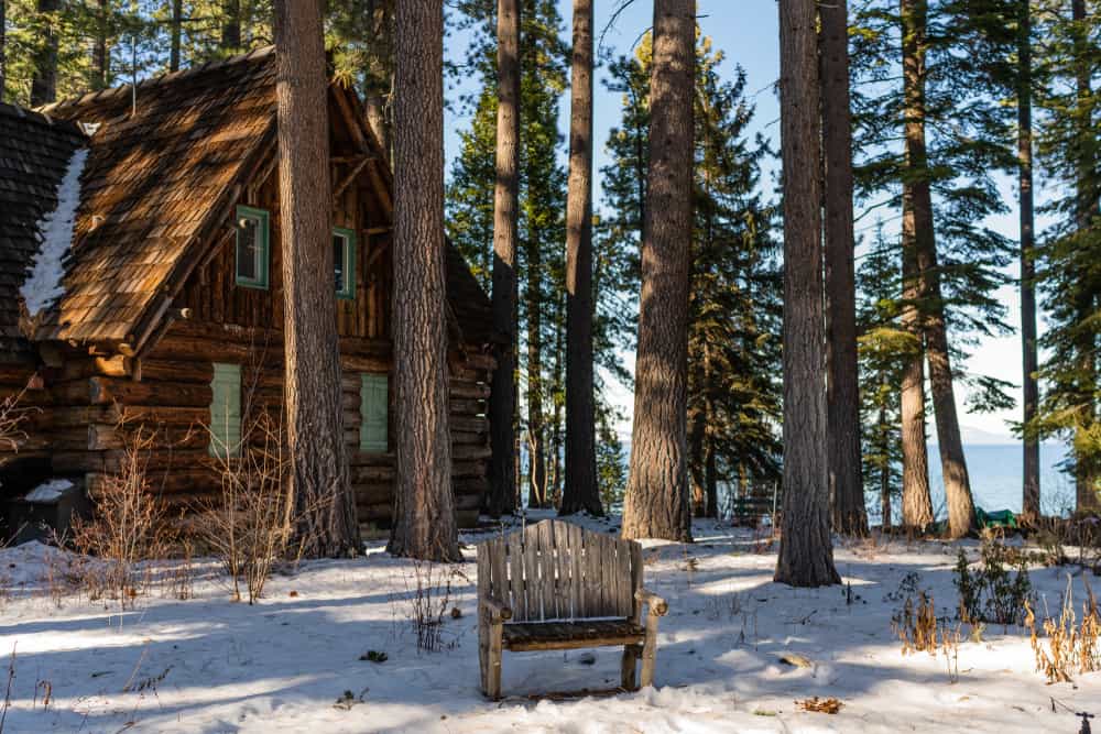 Old wood cabin with a wooden chair in the center of the photo next to a lake with some light snow on the ground.